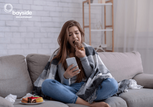 young girl with long hair, wearing jeans and covered with a blanket, sitting with her legs crossed on a couch, looking at a silver colour mobile phone and eating a chocolate eclair, with some paper napkins and a plate full of cakes on her side and the Bayside Psychotherapy logo on the top left