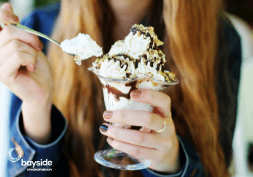 beautifully manicured hand holding a big glass cup with icecream covered with creme chantilly and chocolate, while the other hand holds a full teaspoon of cream with the blurred copper hair of the girl in the background and the Bayside Psychotherapy logo on the bottom left