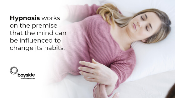 image of a white skin woman with long blond hair and a pink pullover in a hypnosis state and her left hand lifted by another hand, on a white background, with a text about how hypnosis influences the mind and with the Bayside Psychotherapy logo at the bottom left