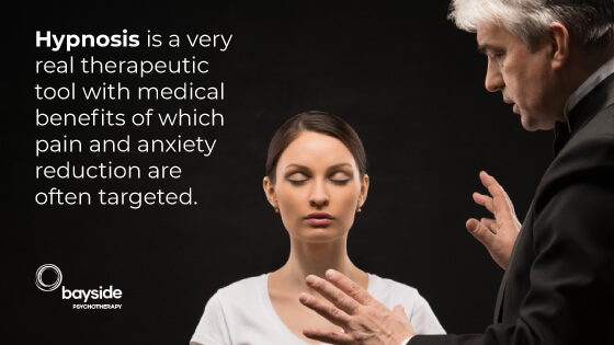image of a white skin woman with a white t-shirt in a hypnosis state and a man with white hair talking to her with a text about hypnosis as a real therapeutic tool and with the Bayside Psychotherapy logo at the bottom left